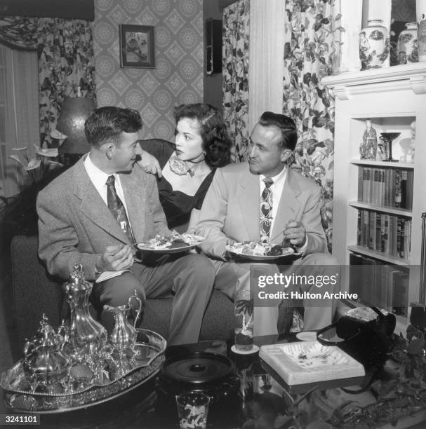 Two male hypnotists sit and eat on a living room sofa as a woman leans down between them, staring at one of the men, at a party for hypnotists.