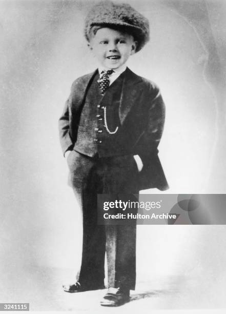 Full-length portrait of American actor Mickey Rooney as a young boy, wearing a three-piece suit, tie, and large cap. Rooney is smiling and has his...