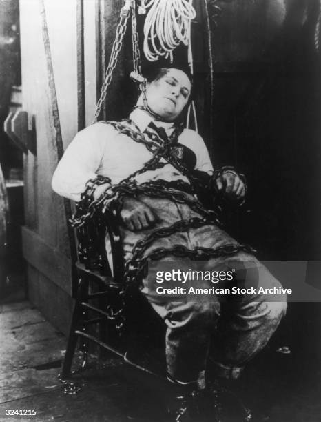 Hungarian-born magician Harry Houdini is bound to a chair with chains and shackles.