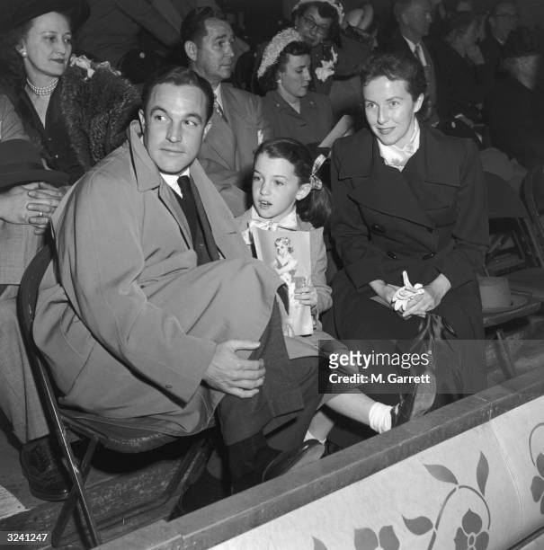 American dancer and actor Gene Kelly , his wife, Betsy Blair, and their daughter, Kerry, holding a program, sit in the stands at an Ice Follies...