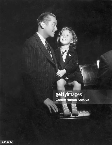 American child actor Shirley Temple sits on a grand piano and smiles at actor James Dunn during rehearsals for their fourth film together, 'Baby Take...