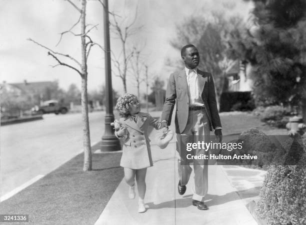 American child actor Shirley Temple holds hands with actor, tap dancer Bill 'Bojangles' Robinson while walking on a sidewalk at 20th Century-Fox...