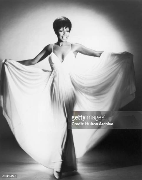 Full-length studio portrait of American actor Diahann Carroll, fanning her long white dress with her arms.