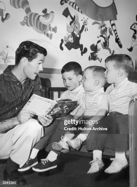 American television host Johnny Carson kneels while he reads a children's book to his sons : Kit, Cory and Ricky, in their nursery. There are cartoon...