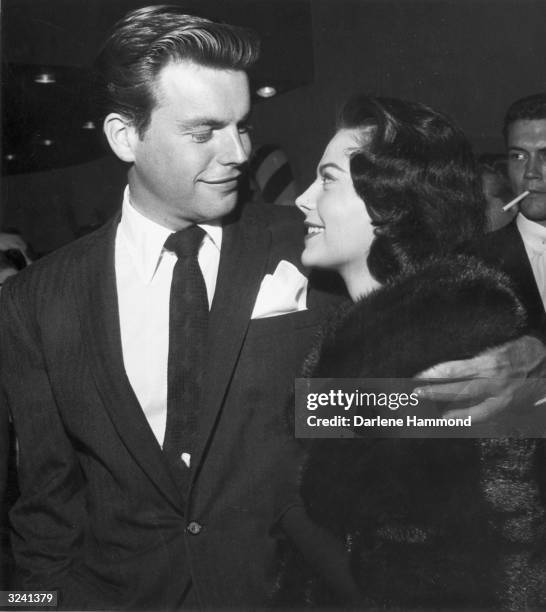 American actor Robert Wagner stands with his arm around American actor Natalie Wood , wearing a fur coat, as the couple looks each other in the eye...