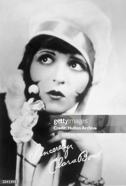 Autographed studio portrait of American actor Clara Bow wearing a cloche hat and a bobbed hairstyle while holding her hand to her face and pursing...