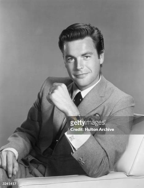 American actor Robert Wagner, wearing a suit and tie with a pocket square, gold watch, and cufflinks, smiling while sitting in an armchair with his...