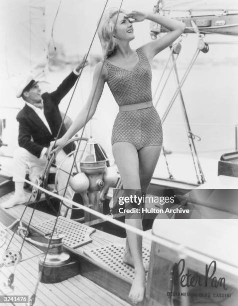 Woman wearing a one-piece 'Peter Pan' bathing suit designed by Oleg Cassini on board a yacht shields her eyes from the sun to look up, as a man...