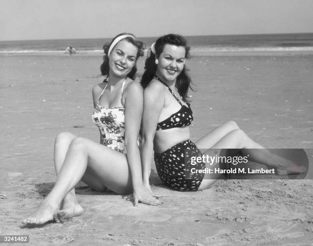 Two women, one wearing a one-piece bathing suit, the other a two-piece, smile as they sit back-to-back on a beach.