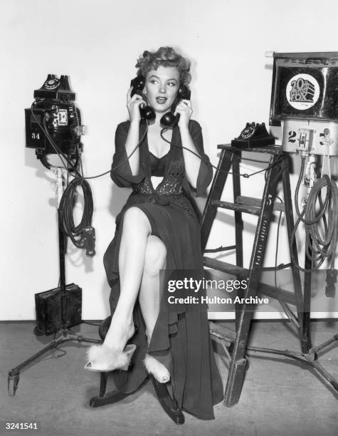 Full-length portrait of American actor Marilyn Monroe holding a telephone receiver to either ear while sitting on a stool in lingerie and...
