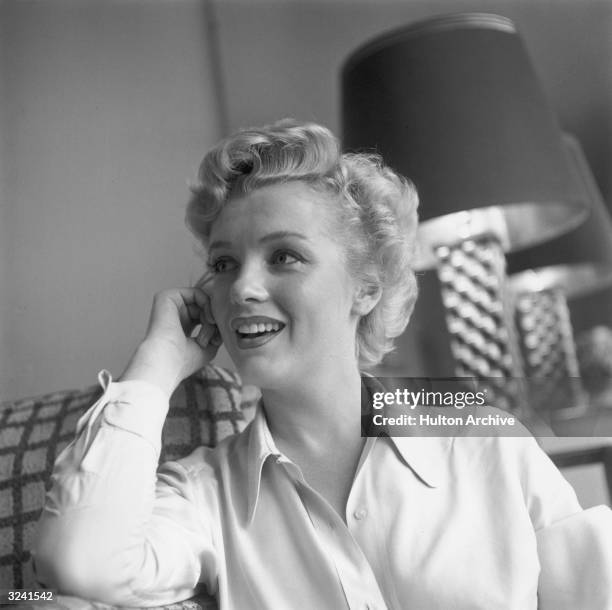 American actor Marilyn Monroe , wearing a button-up long-sleeved shirt, leaning against a chair at her home.