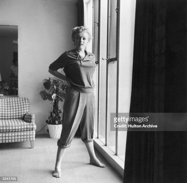 American actor Marilyn Monroe stands barefoot near a window in a sweater and checkered pants.