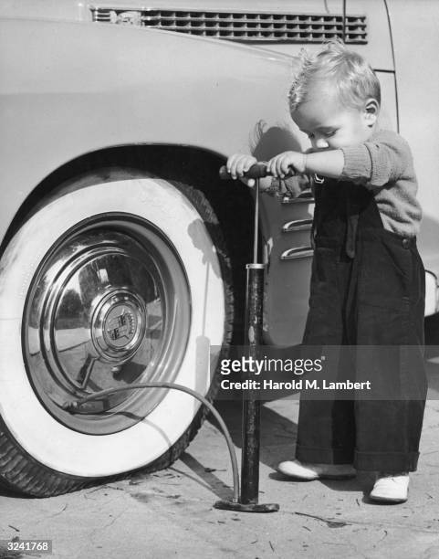 Toddler dressed in overalls attempting to pump air into a car tire, 1940s.