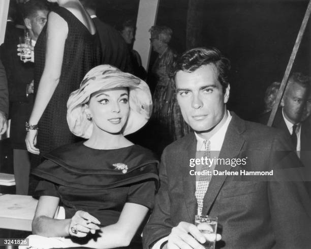 British-born actor Joan Collins smokes a cigarette and American actor and artist Gardner McKay holds a drink as they sit together during a party...