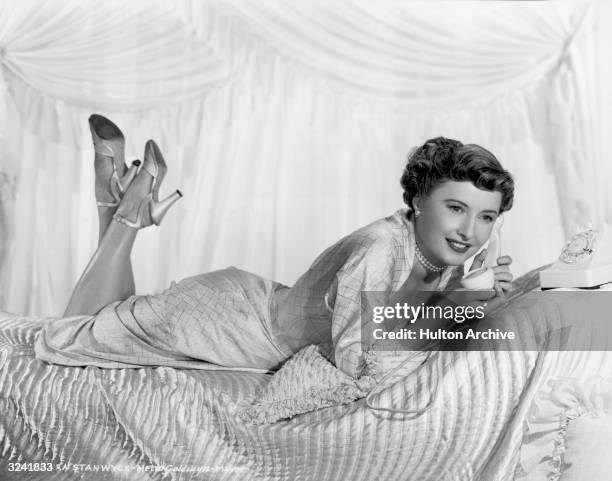 American actor Barbara Stanwyck lies on her stomach with her feet in the air and holds a telephone receiver to her ear while lying on a bed in a...