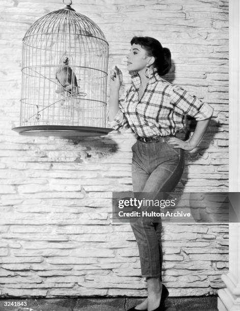 Full-length portrait of British-born actor Joan Collins feeding a parrot in a birdcage a cracker. Collins is wearing jeans and a plaid shirt with the...