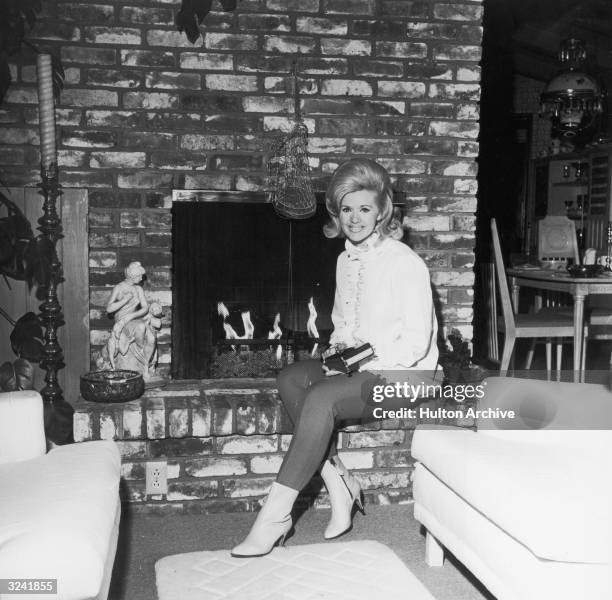 American actor and singer Connie Stevens, wearing a ruffled shirt, dark pants and ankle boots , sits smiling in front of a brick fireplace.
