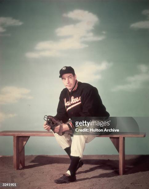 Studio portrait of American baseball player Joe DiMaggio , outfielder for the New York Yankees, sitting on a bench and wearing his uniform in front...