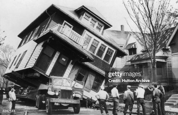 Full-length view of a house tilting off the side of a flatbed truck as a group of movers look on, Wisconsin.