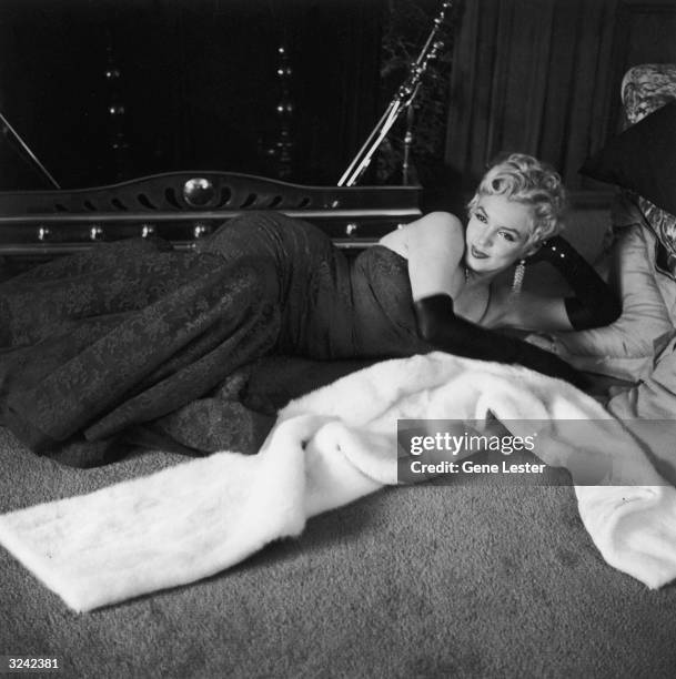 American actor Marilyn Monroe , wearing a lace evening dress and long gloves, lies in front of a fireplace next to her fur wrap.