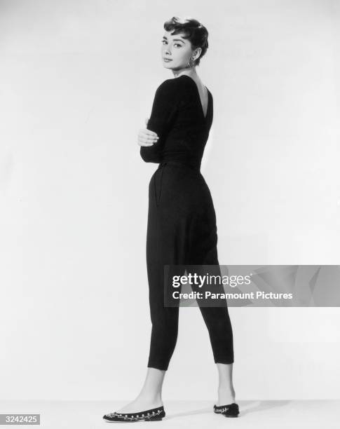 Belgian-born actor Audrey Hepburn, wearing black Capri pants and a black sweater with flats, looks over her shoulder in a full-length promotional...