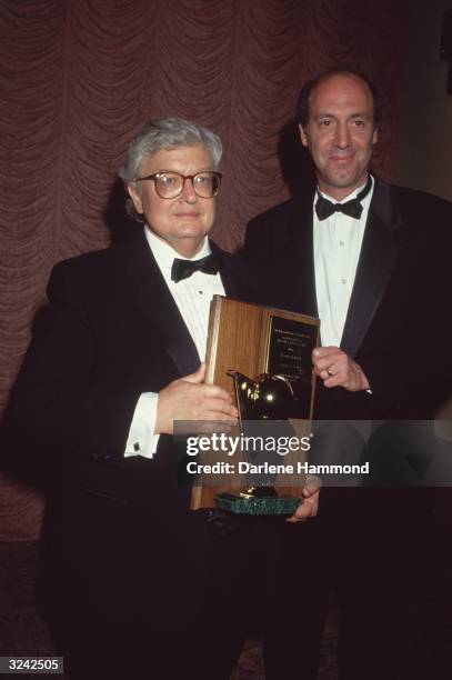 American film critics Roger Ebert and Gene Siskel, hosts of the television series 'Siskel & Ebert at the Movies,' hold an award. They wear tuxedos.