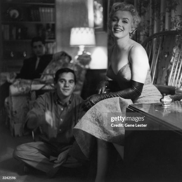 American actor Marilyn Monroe laughs while sitting next to photographer Milton Greene in a living room.