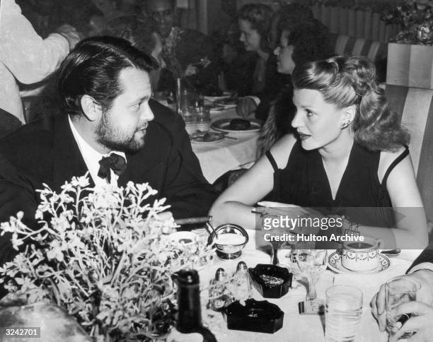 Rita Hayworth and her husband, Orson Welles, sit at a table in a restaurant facing each other while wearing evening clothes.