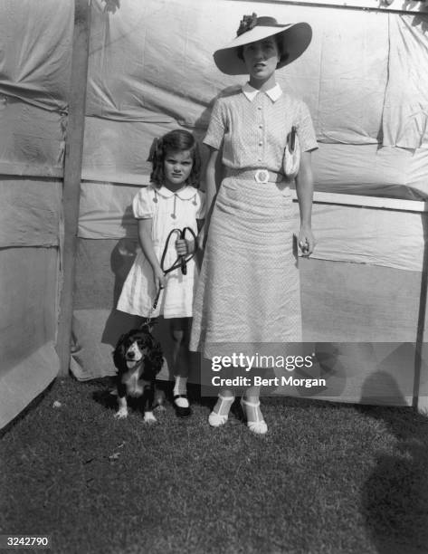 Full-length portrait of Jacqueline Bouvier standing beside her mother, Mrs. John V. Bouvier III, while holding her dog, 'Bonnett,' by its leash at a...