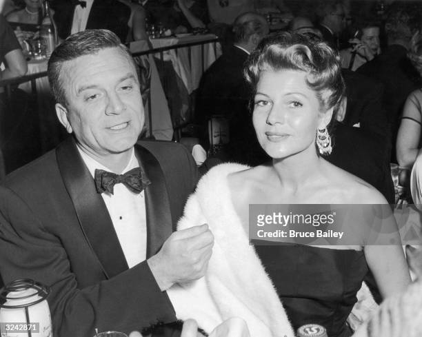 American actor Rita Hayworth , wearing an off-the-shoulder gown with a white ermine fur wrap, sits next to her fourth husband, James Hill, at a table...