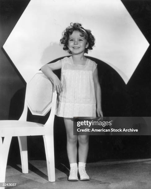 Full-length portrait of child actor Shirley Temple wearing a short dress, standing and smiling as she poses with her arm on the back of a chair.