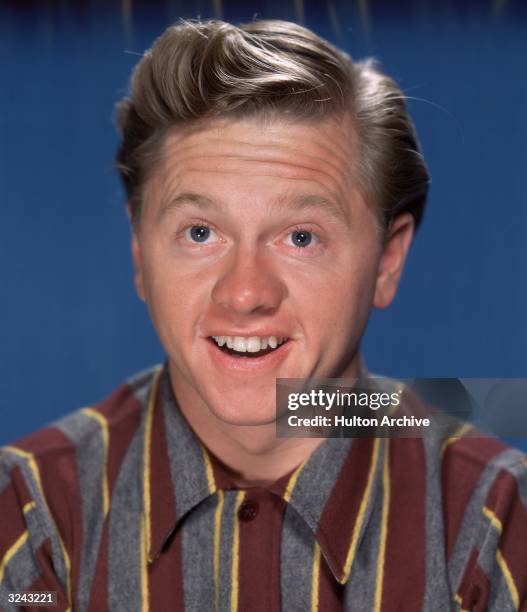 Promotional portrait of American actor Mickey Rooney, smiling broadly in front of a blue backdrop, circa 1955. He wears a striped flannel shirt.