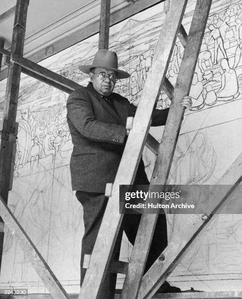 Mexican painter Diego Rivera stands on a wooden platform, holding a ladder in front of his mural in progress entitled, 'The Great City of...