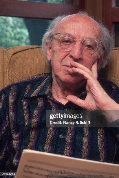 Headshot portrait of American composer Aaron Copland sitting and holding a musical score with his hand up to his face.