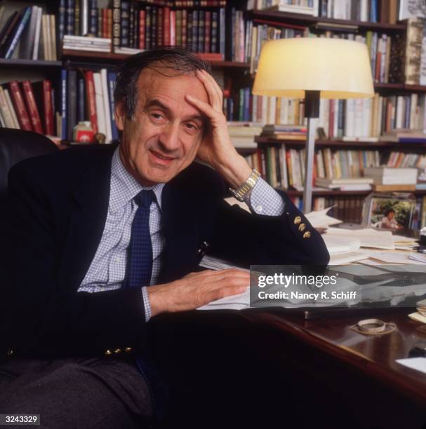 Portrait of Romanian-born author and concentration camp survivor Elie Wiesel sitting at a desk in front of bookshelves, leaning his head on one hand.
