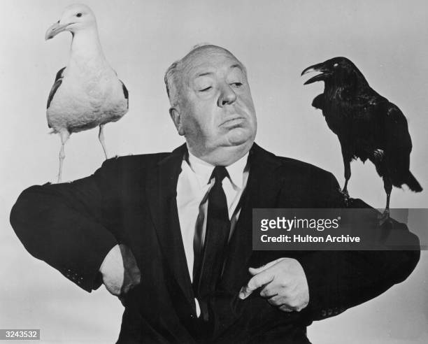 British film director Alfred Hitchcock poses with a seagull and a raven perched on either arm in a promotional still for his film, 'The Birds'.