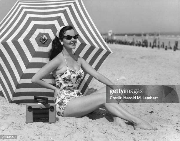 Swimsuit-clad woman wearing cat-eye sunglasses poses on a beach with a radio and a striped beach umbrella.