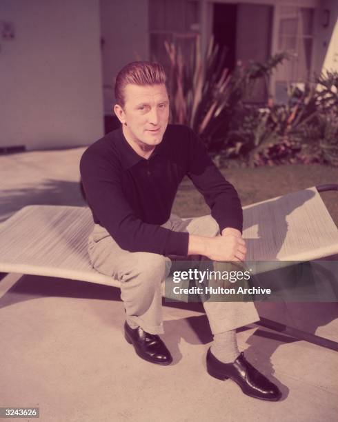 Portrait of American actor Kirk Douglas sitting on a linen sun lounger on an outdoor patio, 1950s. He holds his hands clasped over one knee.