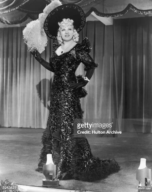 Full-length portrait of American actor Mae West standing on stage wearing a long ruffled and sequined dress and a plumed hat, from director Edward...