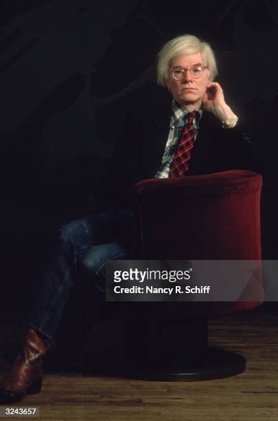 Portrait of American artist Andy Warhol sitting in a red velvet chair against a black background with his hand up to his face. He wears a jacket and...