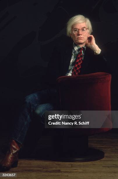 American artist Andy Warhol sitting in a red velvet chair against a black background with his hand up to his face. He wears a jacket and a tie with a...