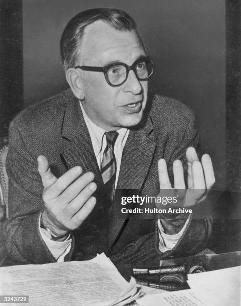 American architect Eero Saarinen, wearing eyeglasses, gestures with his hands while talking in front of a desk.