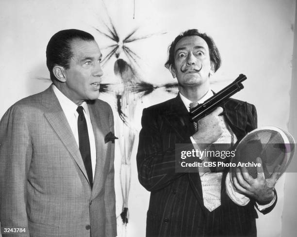 Spanish artist Salvador Dali holds a tranquilizer gun and a fencing mask while posing with American television show host Ed Sullivan in a promotional...