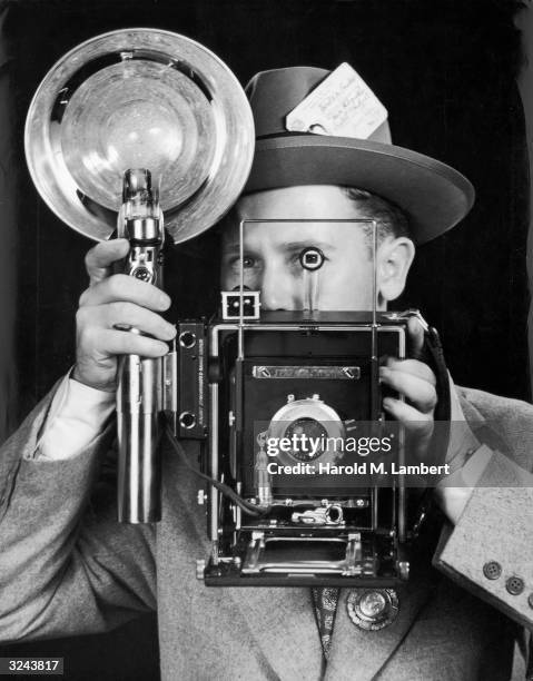 Press photographer looks through the viewfinder of a camera with a flash attached, 1950s.