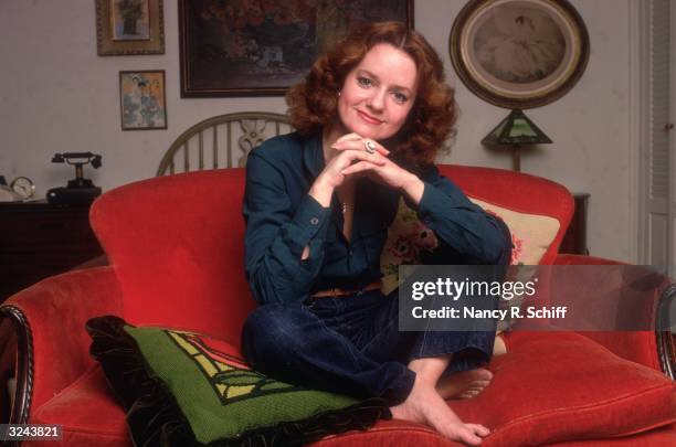 Portrait of American actor Swoosie Kurtz sitting on a red sofa with her legs crossed and her chin resting on her folded hands. She wears jeans with...