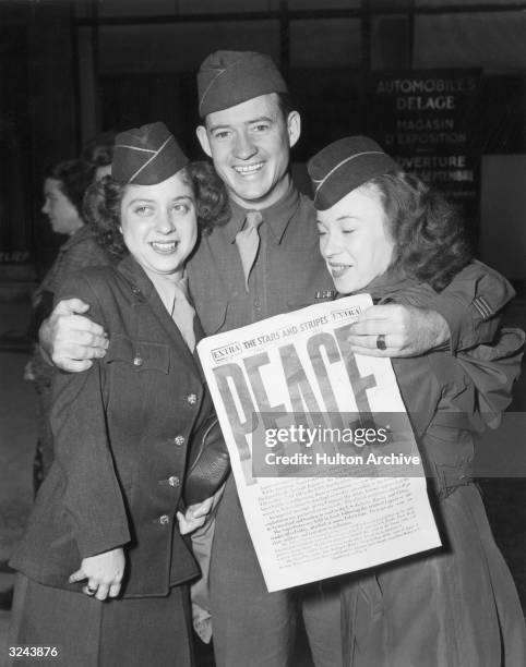 Two women and a man wearing military uniforms smile and embrace as they celebrate the Japanese surrender at the end of World War II. The man holds a...