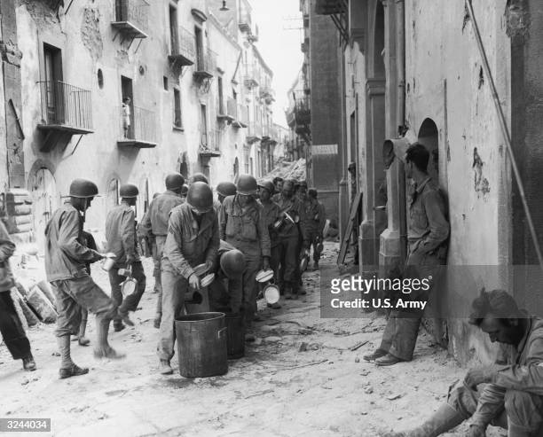 Men of the first US infantry division line up in the street, holding utensils as they wait to eat a meal from a large pot, Troina, Sicily. The Allied...