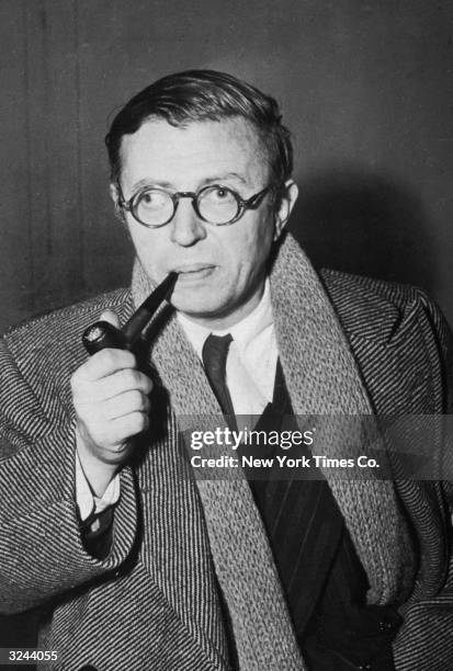 French existentialist author Jean-Paul Sartre smokes a pipe wearing a trenchcoat, scarf and glasses.