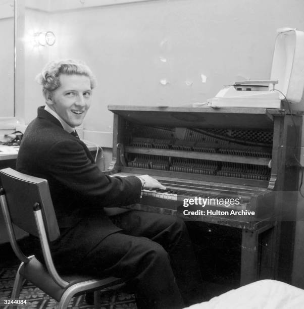 American rock n' roll singer and pianist Jerry Lee Lewis smiles over his shoulder while playing the piano in a dressing room.