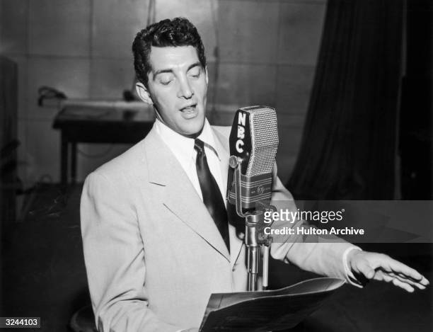 American actor and singer Dean Martin stands behind a microphone, looking at a music stand and singing with one hand out inside the NBC radio studios.
